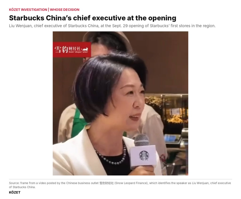 A woman in a white jacket and pearl necklace speaks into a microphone bearing the Starbucks logo, with a Starbucks employee in a green apron behind her.